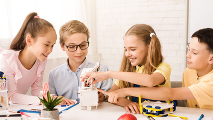 This image shows four children working together on a STEM project in a classroom setting. The children appear to be focused and engaged in their work, and they are all smiling