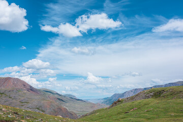 Fototapeta premium Beautiful top view from green pass to wide alpine valley between big mountain range and multicolor mountains under clouds in blue sky. View from above to large valley in sunlight in changeable weather