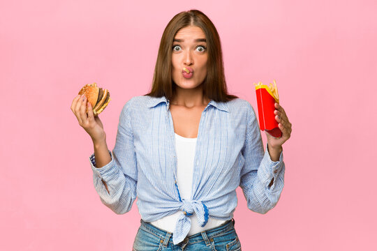 Oops. Surprised Woman Eating Unhealthy Burger And French Fries Standing On Yellow Background. Studio Shot