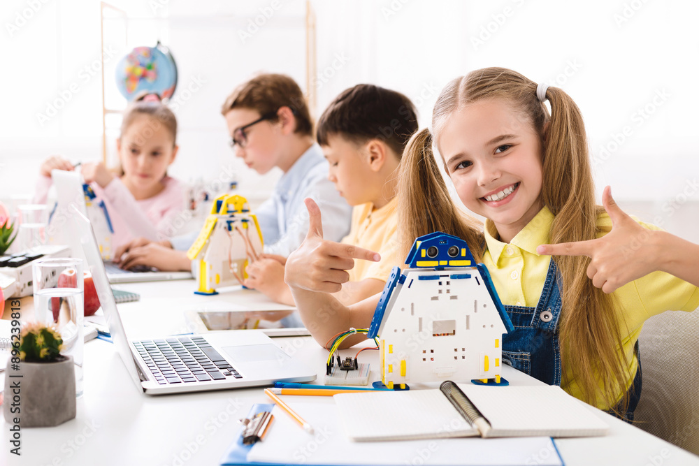 Aufkleber A young girl smiles proudly as she points to a robot she built in a STEM classroom setting. The image shows other students working on similar projects and a laptop with a robotic arm on the desk.