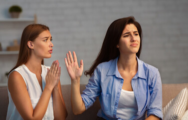 Miserable Girl Asking Indifferent Friend For A Favor Begging Holding Hands In Prayer Gesture Sitting On Sofa Indoor. Selective Focus