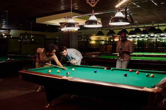 Young man bending over wide pool table while standing next to his girlfriend with cue and teaching her how to play billiards