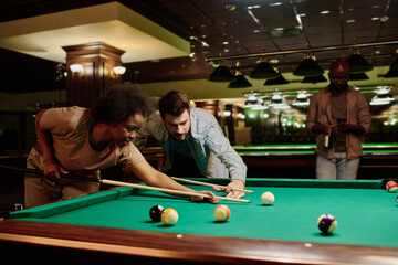 Young male and female billiards players with cues bending over table with green top and getting ready to hit white ball while looking at it