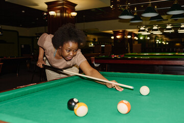 Fototapeta premium Young active woman in casual attire bending over pool table and looking at one of balls before hitting it with cue while playing billiards