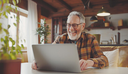 Positive smiling senior grey hair man with glasses communicate with somebody by laptop.He comfortably seating at table in home living room. Active retirement during modern digital world concept.