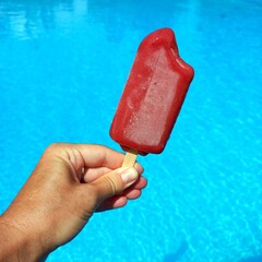 Berry popsicle on a stick in a woman's hand against the background of clear blue water in a swimming pool in the hot summer