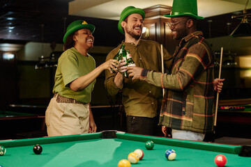 Young excited friends in green hats clinking with bottles of beer standing by wide pool table with balls and celebrating Saint Patrick day