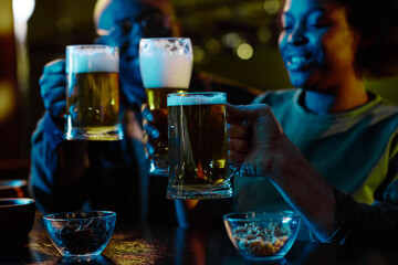 Glass of beer in hand of young unrecognizable man toasting with his friends by bar counter in neon blue lit pub on Saint Patrick day