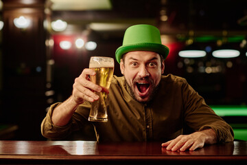 Young excited man in green hat holding glass of beer over bar counter and looking at camera while enjoying celebration of Irish holiday