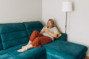 Obraz premium Young woman holding smartphone, using cellphone modern technology, checking cell phone apps, texting, watching video, browsing internet for shopping. Girl sitting on sofa at home. Full length body
