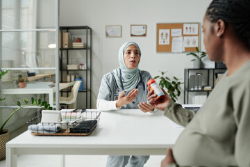 Young female clinician in hijab looking at pregnant woman with bottle of pills or vitamins in hand...