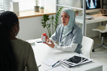 Obraz premium Young confident female clinician in medical scrubs and hijab prescribing pills to African American patient and describing its characteristics