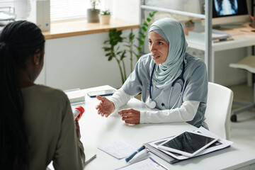 Young Hispanic woman in hijab and medical scrubs looking at female patient with bottle of pills or vitamins during consultation