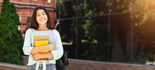 Portraif of smiling asian student girl with backpack and workbooks posing over college campus background, looking at camera, free space