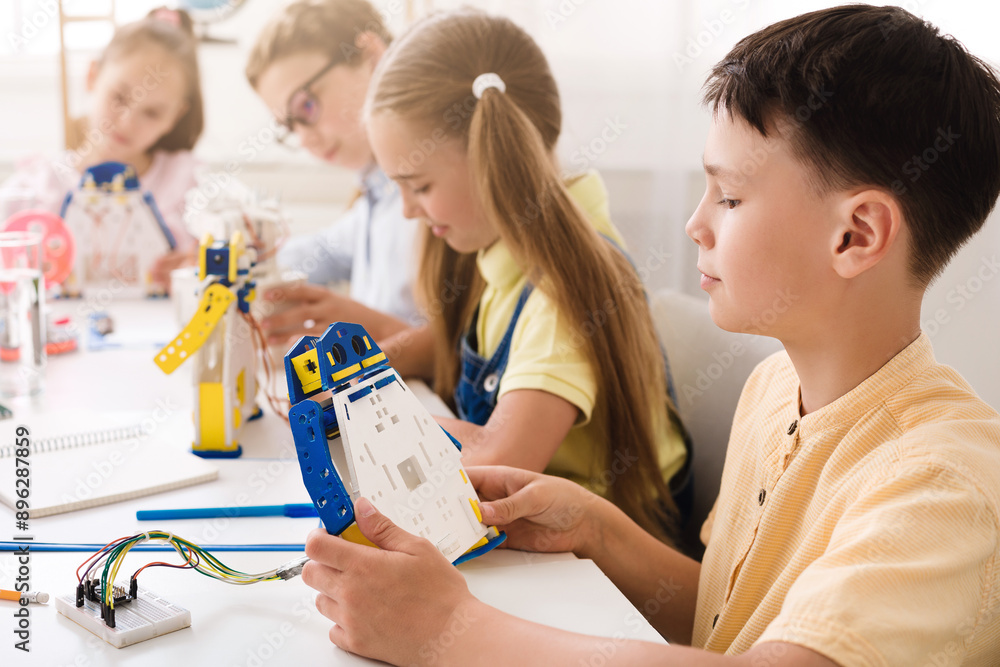 Aufkleber A young boy sits at a table in a classroom and builds a robot with other children. The boy is concentrating on his work as he holds a piece of the robot and examines it.