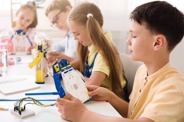 A young boy sits at a table in a classroom and builds a robot with other children. The boy is concentrating on his work as he holds a piece of the robot and examines it.