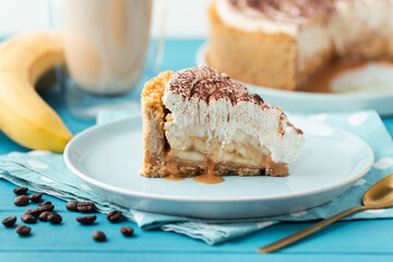 Slice of banoffe pie filled with banana and dulce de leche and sprinkled with grated chocolate. Banana cake on white plate with glass of coffee on turquoise summer background