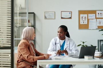 Young African American female clinician in lab coat talking to Muslim patient in hijab and casual...