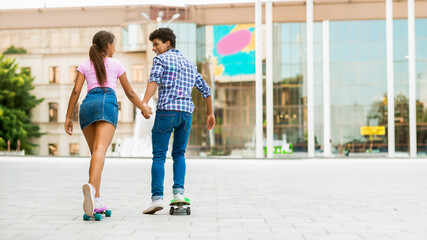 A teen black couple, a girl in a pink shirt and blue skirt, and a boy in a plaid shirt and blue jeans, skateboard side by side in a modern city setting. They are holding hands, copy space