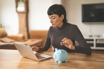 Young Woman Putting Money in Piggy Bank While Using Laptop at Home