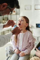 Hands of mature male pediatrician with steel medical spatula checking throat of cute little patient...