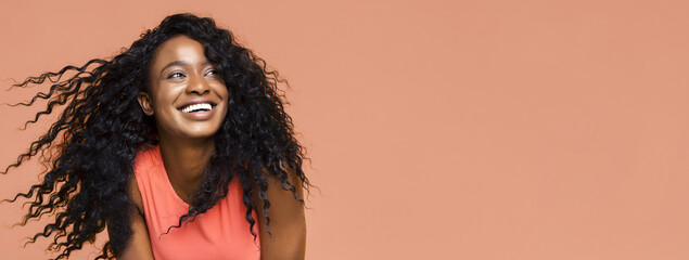 Black woman with curly black hair, wearing a coral sleeveless shirt, smiling and looking off to the...