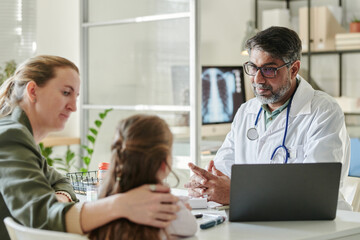 Mature male radiologist sitting by workplace in front of young woman and her little daughter and listening to adorable patient