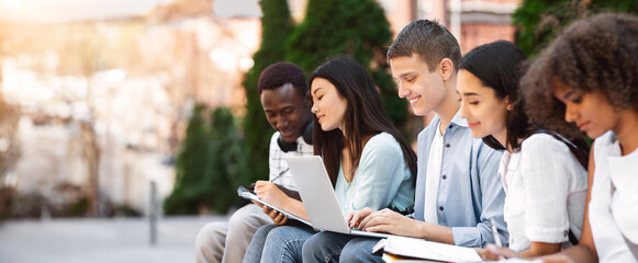 Multiethnic students preparing for lessons outdoors, sitting on bench in campus with workbooks and laptop