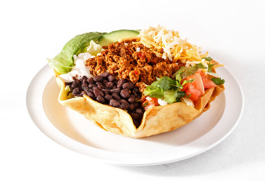 Fresh and healthy taco salad in a flour tortilla shell with black beans, avocado, ground chicken, lettuce, tomato, and cheese in a white plate on a white background; copy space
