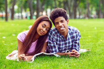 A teen black couple smiles at the camera while lying on a blanket in a grassy park. The girl is resting her head on guys shoulder. They are enjoying a sunny summer day together.