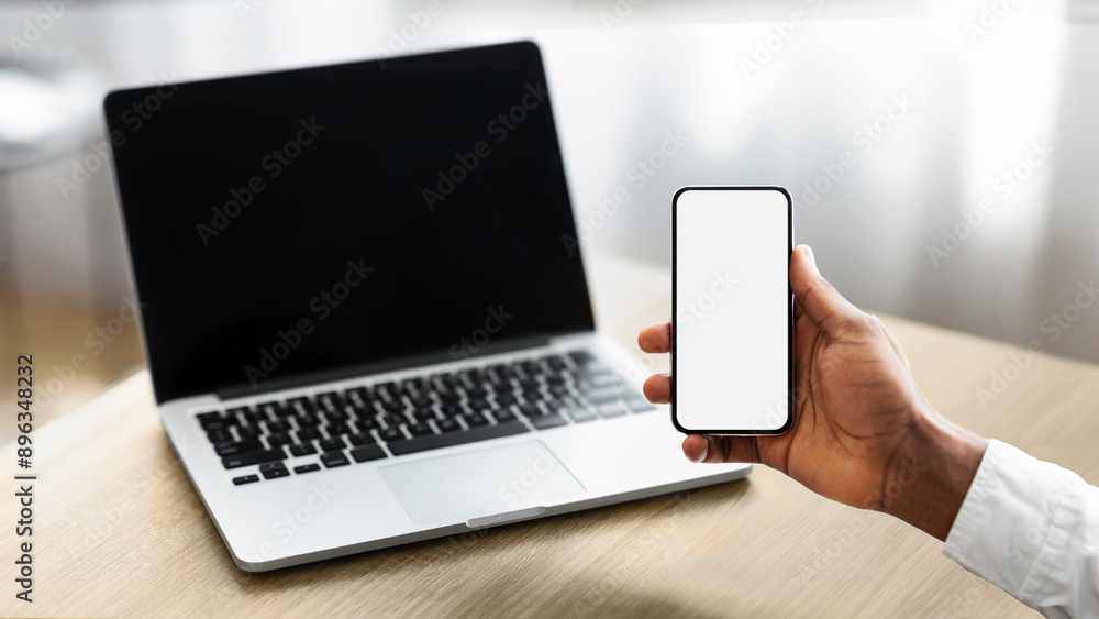 Poster unrecognizable african american businessman holding smartphone with blank screen and working on lapt