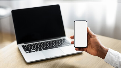 Unrecognizable african american businessman holding smartphone with blank screen and working on laptop in modern office, closeup with copy space