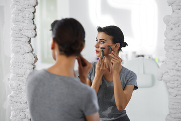 A young woman massages her face with gouache while looking in the mirror.