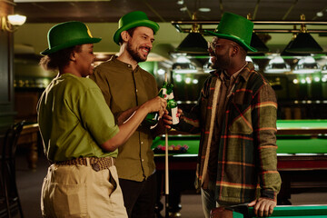 Smiling intercultural friendly people wearing casual attire and green hats looking at each other and toasting with beer during celebration