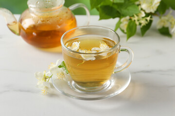 Aromatic jasmine tea in cup, flowers, teapot and green leaves on white marble table
