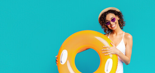African American woman with curly brown hair and a straw hat is smiling and holding a large yellow inflatable ring in front of a blue background. She is wearing purple sunglasses and a white swimsuit