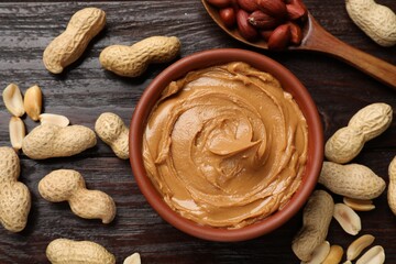 Tasty peanut butter in bowl and nuts on wooden table, flat lay