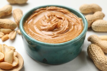Tasty peanut butter in bowl and groundnuts on white table, closeup