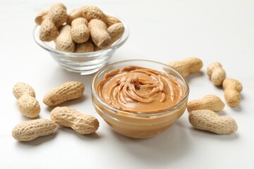 Tasty peanut butter in bowls and groundnuts on white table, closeup
