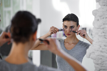 A young woman massages her face with gouache while looking in the mirror.