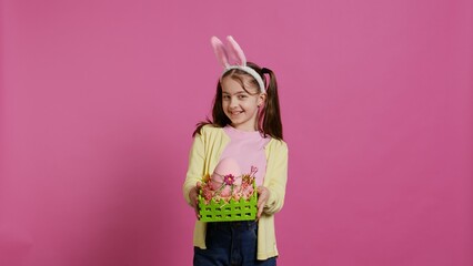 Smiling pleased girl presenting her handcrafted easter basket filled with painted eggs and other festive decorations for holiday celebration. Joyful small kid proud of her arrangement. Camera B.