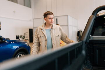 Young man buyer looking around big open trunk of car. Male customer choosing pickup truck in car dealership. Guy visitor walking in modern auto showroom and choosing new vehicle to buy or rent.