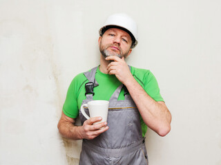 A man in a green shirt and gray overalls holding a cup of tea or coffee. Male construction worker...