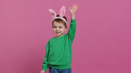 Joyful small kid waving and greeting someone in studio, smiling against pink background. Lovely little boy wearing fluffy bunny ears on his head, celebrating easter holiday. Camera A.