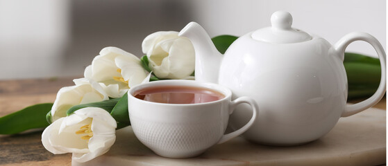 Board with hot brewed cup of tea, pot and tulips on table against blurred background