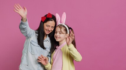Smiling cheerful mother and little girl waving in front of camera, having fun and enjoying easter holiday celebration. Joyful schoolgirl posing with her mom in studio, saying hello. Camera A.