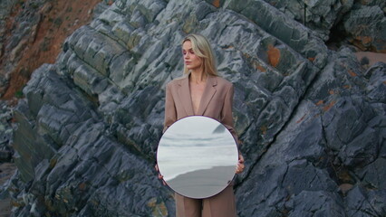 Seashore girl posing rocks zoom on. Serious attractive lady holding round mirror