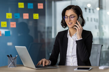 Confident businesswoman multitasking in modern office environment. Engaged in phone call while...