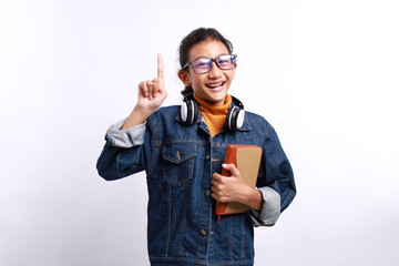 Fototapeta premium Smart kid in casual clothes, headphones and eyeglasses holding book and pointing index finger on white background. Aha moments, getting idea.