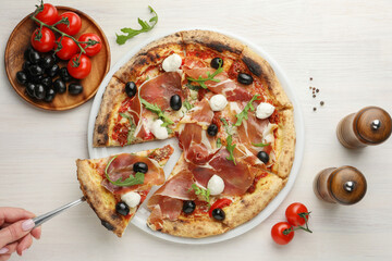 Woman taking piece of tasty pizza with cured ham, olives, mozzarella cheese and arugula at light...